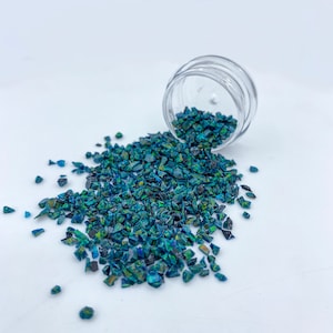 May include: A small clear jar with a lid spills out iridescent green and blue glass shards onto a white surface.