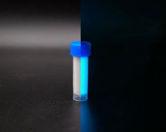 Puede incluir: Un pequeño vial transparente con una tapa azul, medio lleno de una sustancia blanca e iluminado con una luz azul brillante. El vial está centrado contra un fondo oscuro, con la luz proyectando un brillo azul.
