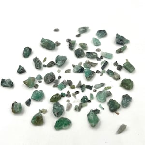 May include: A collection of small, rough-cut emerald gemstones scattered on a white surface. The stones vary in size and shape, displaying shades of green and gray. Some have a crystalline appearance.