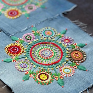 Op de afbeelding: Een close-up van een denim stof met kleurrijke geborduurde cirkels en bloemen. De cirkels zijn gemaakt van verschillende gekleurde draden en hebben een concentrisch ontwerp. De bloemen zijn ook geborduurd met verschillende gekleurde draden en hebben verschillende vormen.