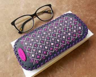 May include: A black perforated leather eyeglass case featuring a geometric cross-stitch pattern in shades of pink and purple. The case has a purple stitched border and a pink oval tag with the word HandMade embossed on the front surface.