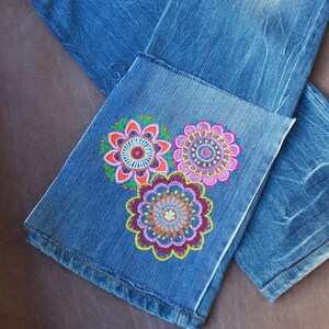 May include: Blue denim jeans featuring a hand-painted design on the lower leg. The artwork consists of three circular floral patterns in vibrant shades of pink, orange, yellow, purple, and green, arranged in a cluster on the fabric surface.