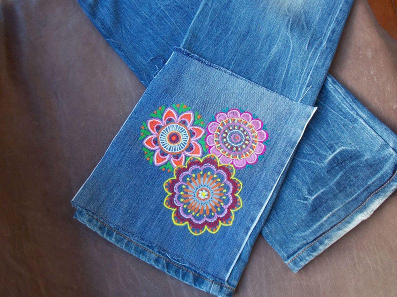May include: Blue denim jeans featuring a hand-painted design on the lower leg. The artwork consists of three circular floral patterns in vibrant shades of pink, orange, yellow, purple, and green, arranged in a cluster on the fabric surface.