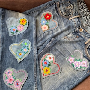 May include: Blue denim jeans featuring five heart-shaped patches. Each patch is made of light blue denim and decorated with colourful floral embroidery in shades of red, yellow, pink, blue, and white. The jeans have a classic five-pocket design.