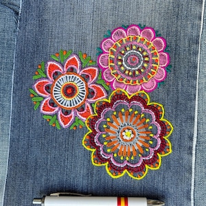 May include: A square patch of blue denim fabric featuring three circular, multi-colored floral embroidery designs in pink, orange, red, and green. A white ballpoint pen with a small Spanish flag detail is positioned horizontally below the embroidery.