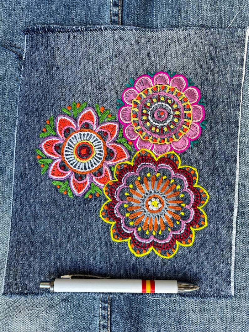 May include: A square patch of blue denim fabric featuring three circular, multi-colored floral embroidery designs in pink, orange, red, and green. A white ballpoint pen with a small Spanish flag detail is positioned horizontally below the embroidery.