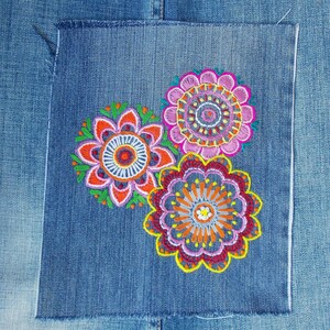 May include: A square denim patch featuring three circular floral embroidery designs in vibrant shades of pink, orange, yellow, and green. The embroidery displays intricate concentric patterns and scalloped edges, stitched onto a blue denim fabric.