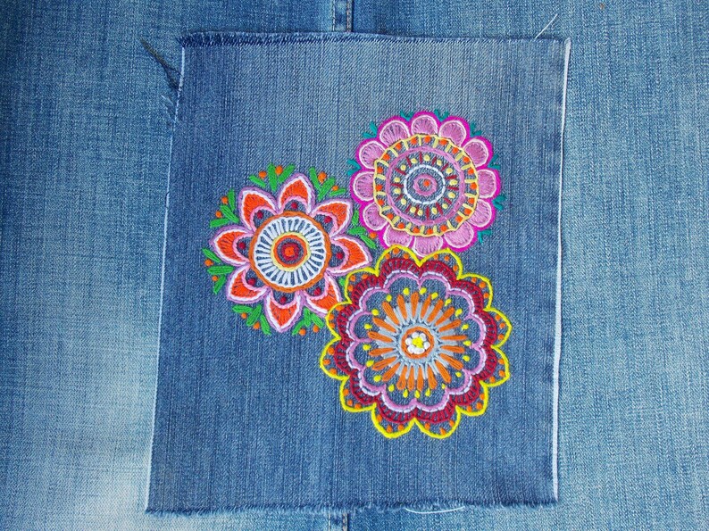 May include: A square denim patch featuring three circular floral embroidery designs in vibrant shades of pink, orange, yellow, and green. The embroidery displays intricate concentric patterns and scalloped edges, stitched onto a blue denim fabric.