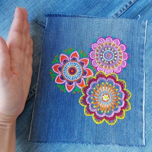 May include: A rectangular piece of blue denim fabric featuring three embroidered floral mandala designs in vibrant shades of pink, orange, yellow, purple, and green. The embroidery is detailed with intricate circular patterns and scalloped edges.