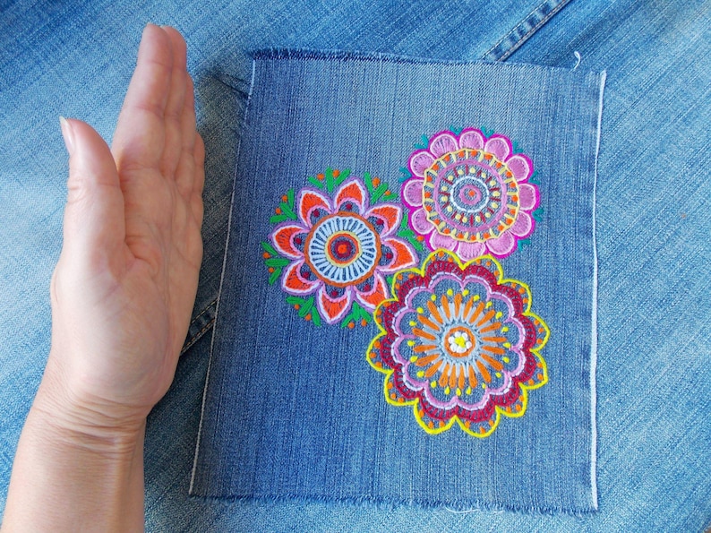 May include: A rectangular piece of blue denim fabric featuring three embroidered floral mandala designs in vibrant shades of pink, orange, yellow, purple, and green. The embroidery is detailed with intricate circular patterns and scalloped edges.