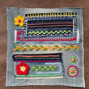 Può includere: Un'opera d'arte tessile in denim con ricami e dettagli all'uncinetto colorati. Include un fiore all'uncinetto giallo, un fiore all'uncinetto rosso e rosa e motivi geometrici ricamati in vari colori. Sono presenti anche due cerchi ricamati.