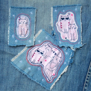 Könnte beinhalten: Drei gestickte Aufnäher mit rosa Katzen mit schwarzen Augen und weißen Flecken auf einem blauen Jeansstoff. Ein Aufnäher zeigt eine einzelne Katze, ein anderer zeigt zwei Katzen und der dritte zeigt drei Katzen.