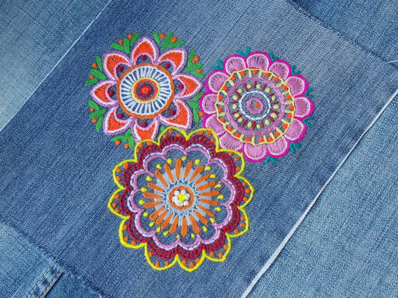 The image shows a detailed embroidery of colorful floral patterns on denim fabric. The vibrant hues and intricate designs stand out against the blue background creating a striking and artistic look. The overall effect is both elegant and eye-catching