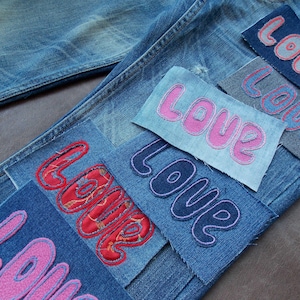 Op de afbeelding: Blauwe spijkerbroek met meerdere rechthoekige stoffen patches op het oppervlak genaaid. Elke patch toont het woord LOVE in verschillende kleuren, waaronder roze, rood en blauw, met geborduurde omlijningen en diverse stoftexturen.