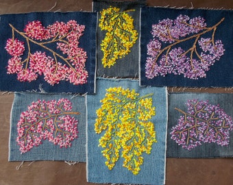 Può includere: Sei toppe rettangolari in tessuto denim con disegni di rami ricamati a mano. Le toppe presentano fiori a punto annodato in varie tonalità di rosa, giallo e viola su sfondi di jeans blu chiaro e scuro con bordi sfilacciati.