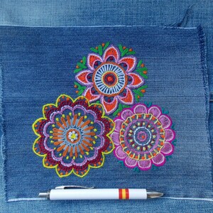 May include: A square patch of blue denim fabric featuring three circular, multi-colored floral embroidery designs in shades of orange, pink, purple, and green. A white pen with a red and yellow striped accent lies horizontally below the embroidery.