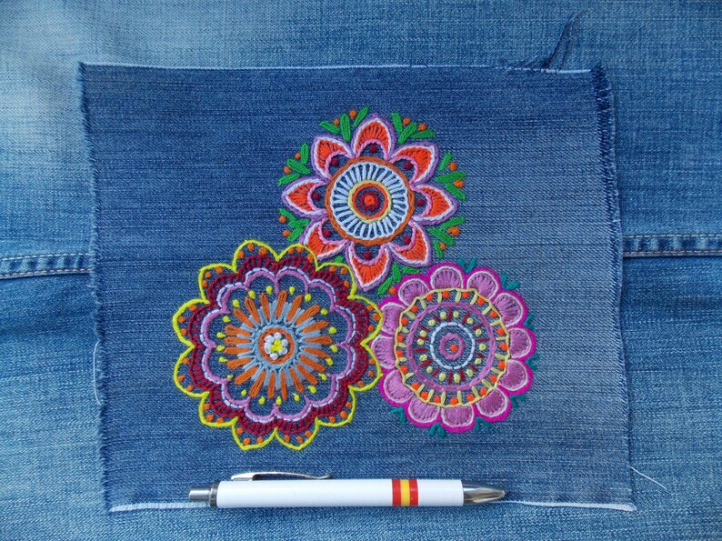 May include: A square patch of blue denim fabric featuring three circular, multi-colored floral embroidery designs in shades of orange, pink, purple, and green. A white pen with a red and yellow striped accent lies horizontally below the embroidery.