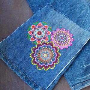 May include: Blue denim jeans featuring three embroidered circular floral mandala patterns on the lower leg. The embroidery uses vibrant threads in shades of pink, orange, yellow, purple, and green, creating intricate, symmetrical, and colorful designs.