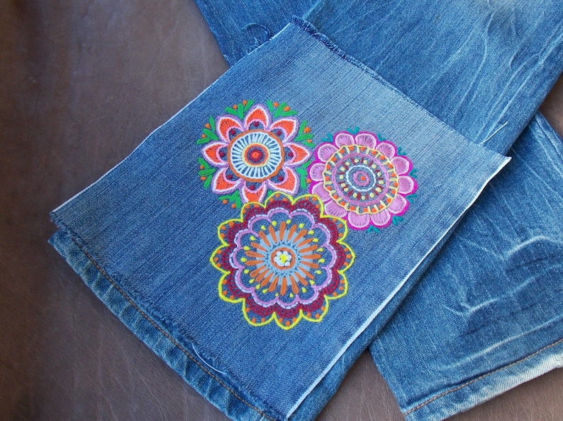 May include: Blue denim jeans featuring three embroidered circular floral mandala patterns on the lower leg. The embroidery uses vibrant threads in shades of pink, orange, yellow, purple, and green, creating intricate, symmetrical, and colorful designs.
