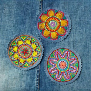 Op de afbeelding: Drie kleurrijke, geborduurde bloemvormige patches met geschulpte randen worden getoond op een blauwe denim achtergrond. De bloemen hebben ingewikkelde ontwerpen in tinten geel, oranje, rood, groen en blauw, waardoor een levendige, handgemaakte uitstraling ontstaat.