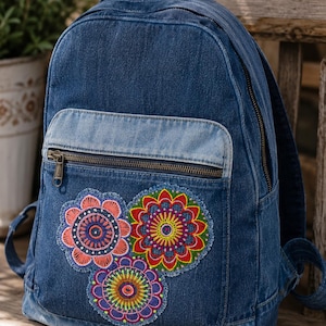 May include: A blue denim backpack featuring a front zippered pocket and three embroidered floral mandala patches in shades of pink, red, yellow, green, and purple. The backpack has a top handle, adjustable shoulder straps, and a brass-toned zipper.