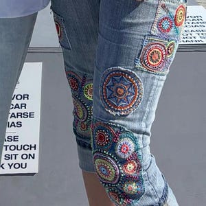 May include: Light blue denim capri pants featuring multiple circular embroidered patches in vibrant colors including red, orange, yellow, green, and blue. The embroidery displays intricate geometric and floral patterns stitched directly onto the fabric.