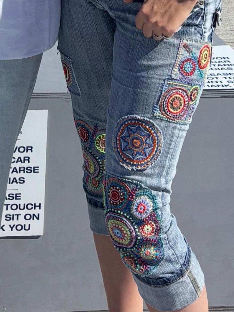 May include: Light blue denim capri pants featuring multiple circular embroidered patches in vibrant colors including red, orange, yellow, green, and blue. The embroidery displays intricate geometric and floral patterns stitched directly onto the fabric.