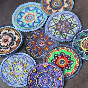 Puede incluir: Una colección de diez posavasos circulares bordados. Cada posavasos presenta un diseño geométrico único y colorido con costuras intrincadas. Los posavasos están hechos de tela vaquera y tienen un borde festoneado. La paleta de colores incluye azules, verdes, naranjas y morados.