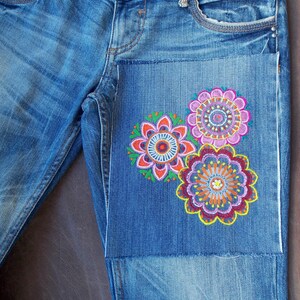 May include: Blue denim jeans featuring a rectangular patch on the upper thigh area. The patch is decorated with three embroidered circular floral patterns in vibrant shades of pink, orange, yellow, purple, and green with intricate stitching.