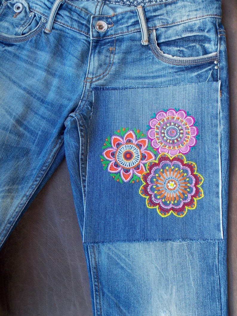 May include: Blue denim jeans featuring a rectangular patch on the upper thigh area. The patch is decorated with three embroidered circular floral patterns in vibrant shades of pink, orange, yellow, purple, and green with intricate stitching.