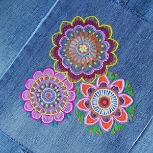 May include: A blue denim fabric featuring three circular embroidered floral designs. The top flower has orange, purple, and yellow petals. The bottom left flower is pink and purple with yellow accents. The bottom right flower is orange and purple.