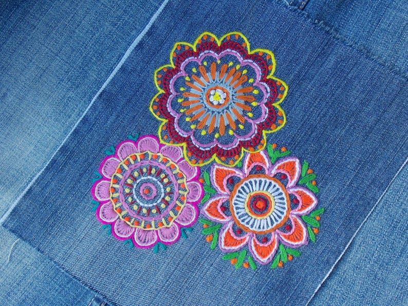 May include: A blue denim fabric featuring three circular embroidered floral designs. The top flower has orange, purple, and yellow petals. The bottom left flower is pink and purple with yellow accents. The bottom right flower is orange and purple.