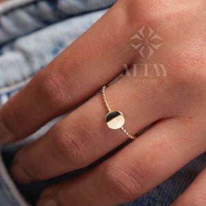 May include: A gold ring with a small, round, flat disc charm. The ring is on a person's finger.