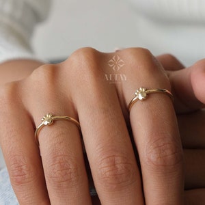 May include: Two delicate gold rings, each featuring a small, flower-shaped design. The rings are displayed on fingers, showcasing their fine details and minimalist style. The rings are a warm gold color.