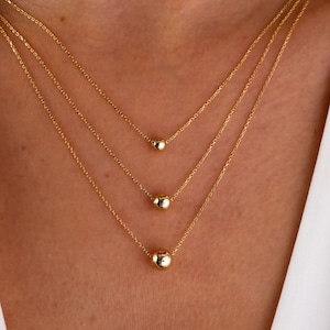 May include: A gold necklace with three graduated sized round beads. The necklace is layered on a model wearing a white shirt.