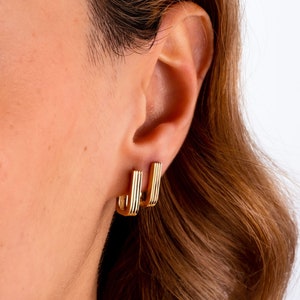 May include: A pair of gold-toned hoop earrings with a ridged design. The earrings are worn in the earlobes of a person with long brown hair.