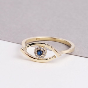 May include: Gold ring with a blue and white eye design. The eye is surrounded by small white stones.