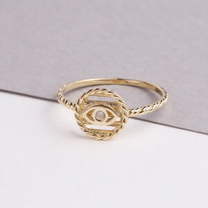 May include: A gold ring with a twisted band and a circular design featuring a stylized eye with a small clear stone in the center.