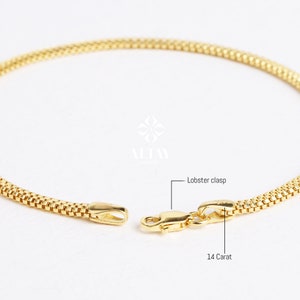 Pode incluir: Close-up de uma pulseira dourada com um design texturizado e tecido. A pulseira apresenta um fecho lagosta e é rotulada como 14 quilates. A joia é exibida em um fundo branco.