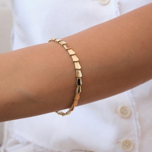 May include: A gold-colored bracelet with a geometric design is displayed on an arm. The bracelet is composed of rectangular segments linked together. The arm is wearing a white shirt with buttons.