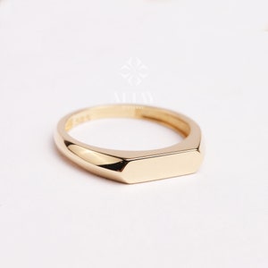 Può includere: Un anello sigillo in oro con una parte superiore piatta e rettangolare. L'anello è semplice ed elegante.