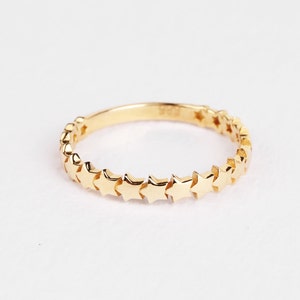 Könnte beinhalten: Ein goldfarbener Ring mit einer durchgehenden Reihe kleiner sternförmiger Verzierungen. Der Ring hat ein glattes, poliertes Band, und die Sterne sind gleichmäßig angeordnet. Der Hintergrund ist schlicht weiß, was das Design des Rings hervorhebt.