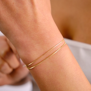May include: A gold chain bracelet with two thin chains layered on top of each other. The bracelet is on a person's wrist.
