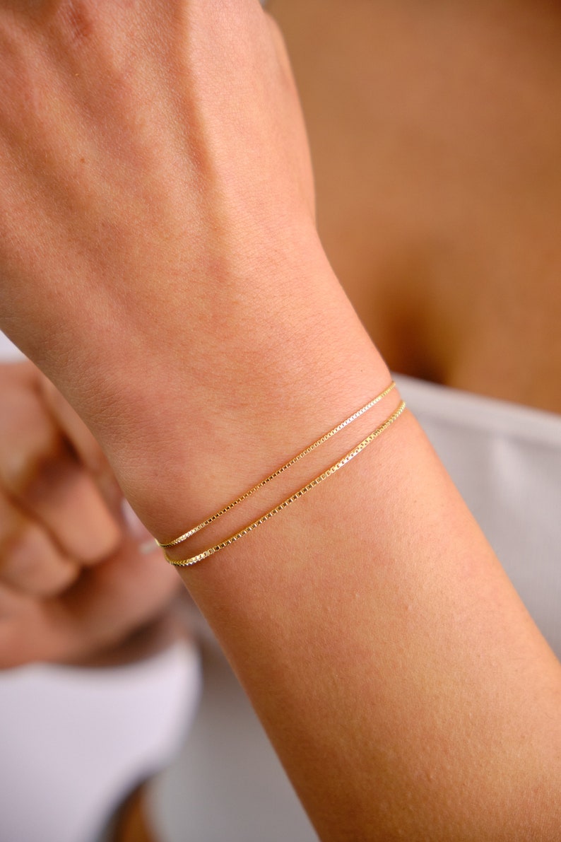 May include: A gold chain bracelet with two thin chains layered on top of each other. The bracelet is on a person's wrist.