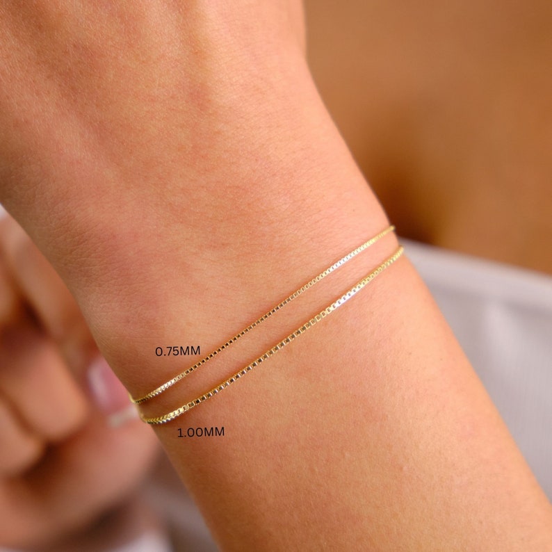 May include: Two gold chain bracelets on a person's wrist. The top bracelet is labelled 0.75MM and the bottom bracelet is labelled 1.00MM.