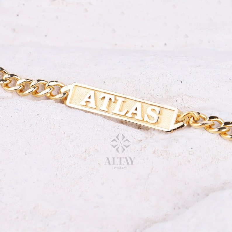 Pode incluir: Um bracelete de corrente de ouro com uma placa retangular que diz "ATLAS" em letras em relevo.