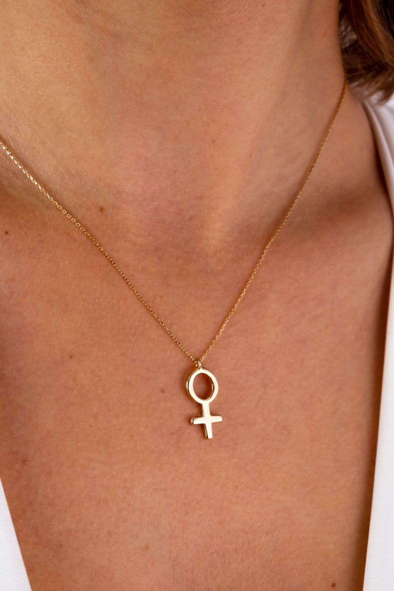 K&ouml;nnte beinhalten: Eine goldene Halskette mit einem schlichten, minimalistischen Anh&auml;nger in Form des weiblichen Symbols.