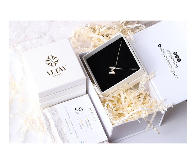 K&ouml;nnte beinhalten: Eine wei&szlig;e Box mit einem goldenen Logo, auf dem "Altay Jewelry" steht, ist ge&ouml;ffnet und zeigt eine wei&szlig;e Box mit einer goldenen Kettenkette mit einem Buchstaben "M" Anh&auml;nger. Die Kette liegt auf einem schwarzen Samtkissen. Die Box ist von wei&szlig;em Knitterpapier umgeben.