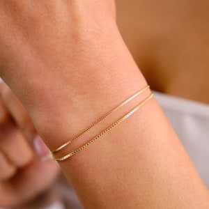 May include: Two gold chain bracelets on a person's wrist. The bracelets are layered, with the thinner chain on top of the thicker chain.
