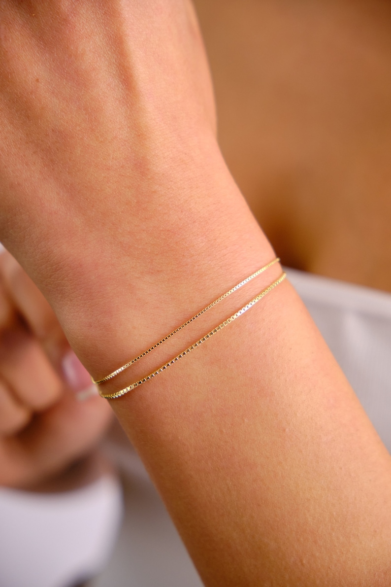 May include: Two gold chain bracelets on a person's wrist. The bracelets are layered, with the thinner chain on top of the thicker chain.